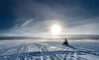 One person drives snowmobile in very cold mountains in Sweden, frosty fog around bright sun creates halo effect, wild birches forest and mountains behind driver. Hemavan -Tarnaby area in Lappland