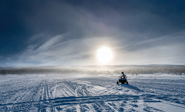 One person drives snowmobile in very cold mountains in Sweden, frosty fog around bright sun creates halo effect, wild birches forest and mountains behind driver. Hemavan -Tarnaby area in Lappland