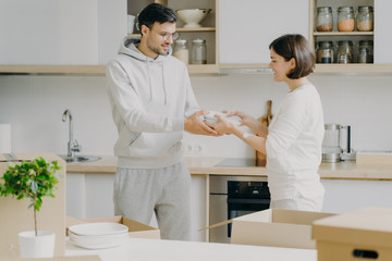 Horizontal shot of smiling couple unpack cardboard boxes in new kitchen, man passes plates to woman, placing dishes in cupboard, surrounded with moving packages in own bought or rented apartment