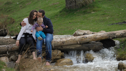 Happy parents with baby child sitting on tree timber over pouring mountain river
