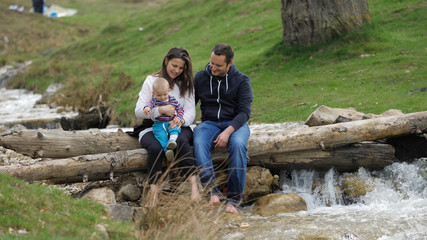 Young mother, father and lovely baby child sit o tree trunk over mountain river