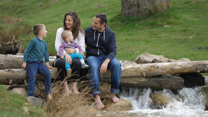 Happy parents, baby and bigger brother sit on tree timber over river, laughing