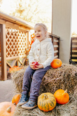 Fototapeta premium Adorable blonde baby toddler in white knittes jacket sitting on the haystack with pumpkins at porch, playing with apple and laughing