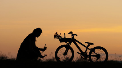 Obraz premium Girl silhouette picking flower on hill near bike, beautiful sunset sky, romantic