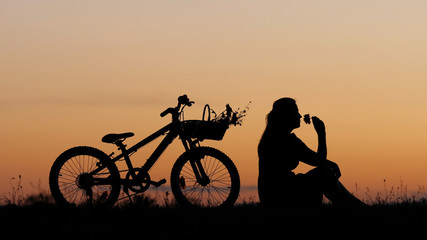 Obraz premium Young woman silhouette sit on hill and play with flower near bike, sunset sky