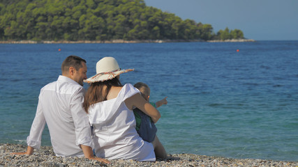 Couple of lovers with child sit on beach and look at blue sea, family holiday