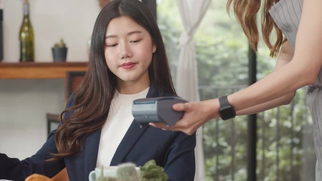 Young Asian business women pay contactless at coffee shop. Asian happy girl barista waiter wear gray apron holding credit card reader machine for customer using mobile phone scan pay in cafe.