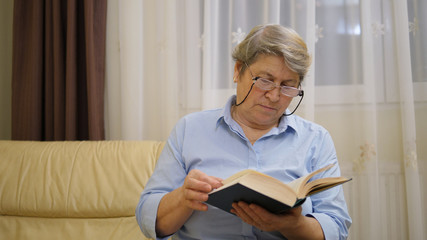 Old woman with gray hair, eyeglasses sit on sofa and read a book