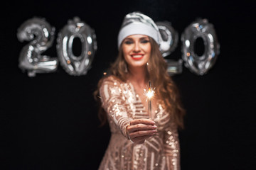 Happy young woman in Santa hat with sparklers in hand against metallic 2020 balloons.
