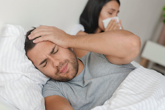 Young couple having flu and lying in bed at home. Man have headache while his wife sneezing at napkin
