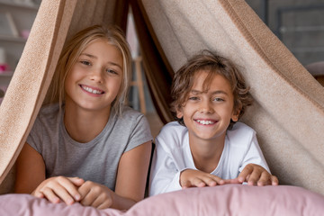 Happy boy and girl sitting in fort from blankets and smiling in camera