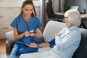 Female nurse checking blood pressure of senior woman at home - obrazy, fototapety, plakaty