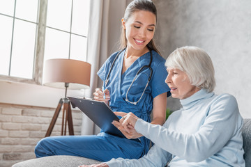 Smiling senior woman reading medical tests results in hands of nurse visiting her