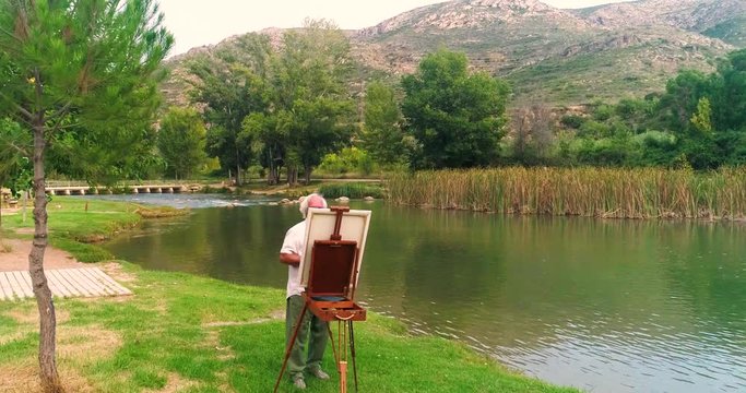Un artista anciano pintando un cuadro en un caballete junto a un río otoñal