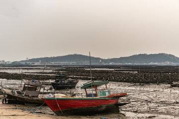 Ban Bai Bua, Thailand - March 16, 2019: Large Oyster beds and banks in the Gulf of Thailand just off shore with forested hills in back. Three stranded sloops during low tide under cloudy sky.