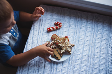 Child boy taking Christmas gingerbread from plate on table near window in daylight.