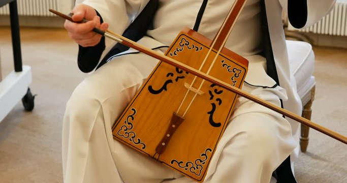 Professional Caucasian impressive musician playing matou qin, chinese mongolian wooden cello. white clothes and beautiful natural light. windows with trees outside