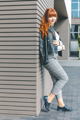 Cute young girl in a black leather jacket with curly red hair and a fringe relaxes near a modern...