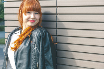 Cute young girl in a black leather jacket with curly red hair and a fringe stands near the modern...