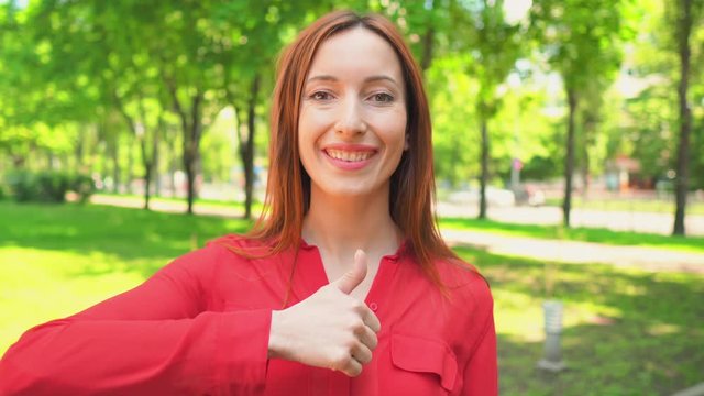 happy caucasian girl showing thumb up hand gesture cheerful female in red shirt standing on the street nature summer background