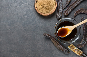 carob molasses in glass bowl and carob powder or flour with carob pods on rustic background, locust...