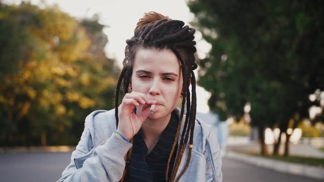 Portrait of young pretty woman with dreadlocks smoking marijuana joint in park, slow motion