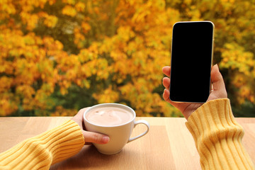 cup of cocoa with milk or cappuccino in one hand and smart phone with blank screen mobile in...