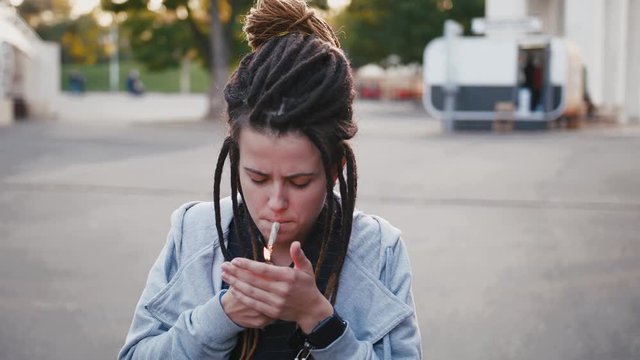 Portrait of young pretty woman with dreadlocks smoking marijuana joint in park, slow motion