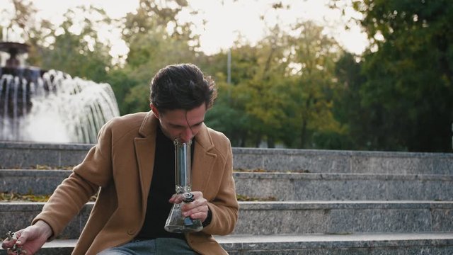 Handsome stylish young man sitting on stairs and smoking weed with bong in park