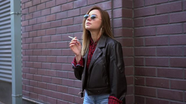 Young Beautiful stylish European girl 30s years dressing black leather jacket jeans and red hat standing on backyard near building and smoking cigarette Stress concept Bad habit unhealthy lifestyle.