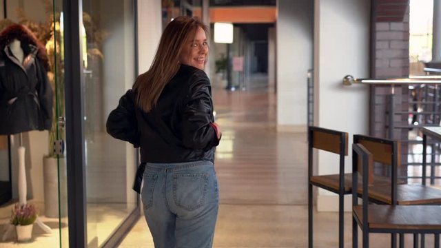 Hand holding shooting A young beautiful Caucasian girl dressed black leather jacket and jeans walks shopping mall turn around and laughs the rear view. modern young adult woman Calling for himself.