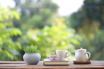 white cup with teapot and small plant with purple daisy flower and book on wooden table