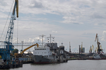 Coal loading or unloading of a grey bulk carrier with a white deck using an excavator in the port, nearby loading platforms and port loading cranes can be seen on the background a cloudy blue sky