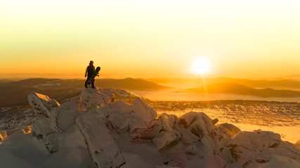 a snowboarder stands on top of a mountain in the Golden rays of the dawn sun. Freerider enjoys the morning sun with a Board in hand standing on large rocks above the clouds. winter activity concept