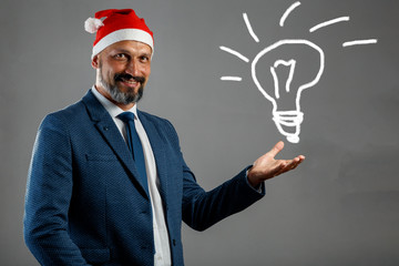 Bearded man in a blue jacket and a Christmas hat on a gray background holding a painted light bulb. Concept of idea for new year, christmas.
