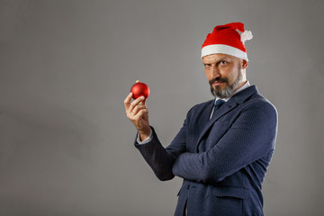 A bearded man in a blue jacket and a New Year's hat on a gray background holding a red Christmas ball for spruce. Concept of young santa claus.