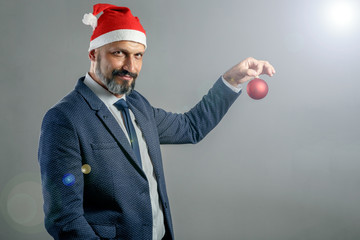 A bearded man in a blue jacket and a New Year's hat on a gray background holding a red Christmas ball for spruce. Concept of young santa claus.