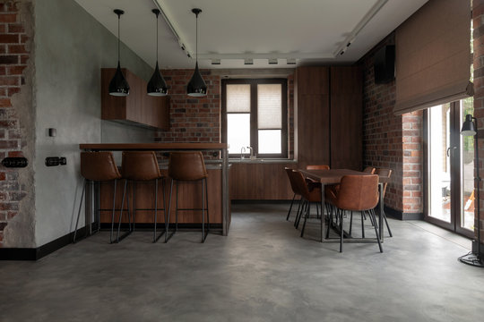 Contemporary interior design of light spacious dinning room including brown wooden furniture with bar stools at counter and soft comfortable chairs at table with cement effect in loft style