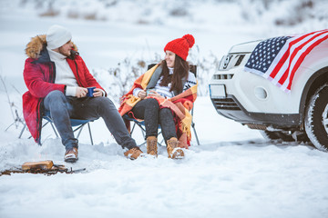 couple sitting in chairs near campfire talking. suv car with usa flag on the hood