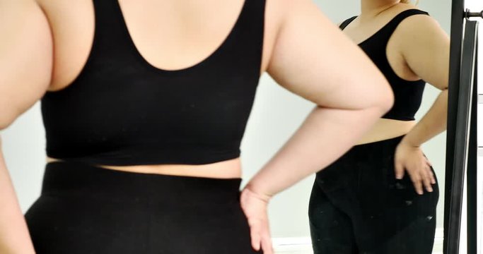Overweight young woman in sportswear looking her belly fat into the mirror