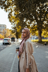 Obraz premium Young business woman of oriental appearance in a beige shawl on her head and a beige cloak stands against the backdrop of a city park and holds a paper cup with coffee in her right hand, smiling