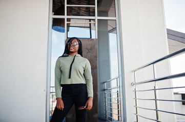 City portrait of positive young dark skinned female wearing green hoody and eyeglasses standing at balkony.