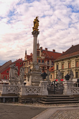 Beautiful landscape view of Plague Column against cloudy sky. Ancient colorful building at the background. The Rotovz Town Hall Square in Maribor, Lower Styria, Slovenia