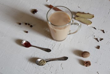 Masala tea in glass cup with spices as fennel seeds and saffron in silver spoons, cinnamon stick, ginger, anis star, nutmeg, pepper, cloves  on wite concrete background