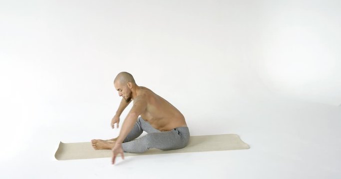 A flexible and plastic male yogi with a beard does yoga on a rug in a White Studio. He is sitting in the butterfly pose-Baddha konasana.