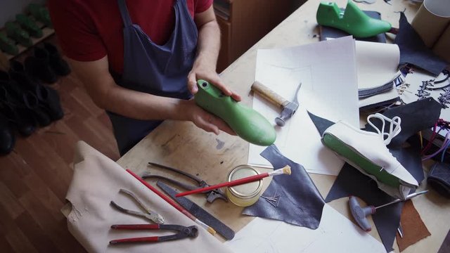 Tracking shot top view of young shoemaker examining plastic last while making white leather sports shoes standing at table in workshop