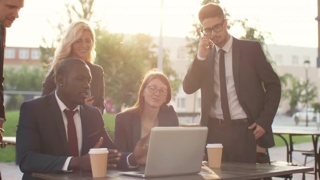 Group of young businesspeople of different ethnicities discussing something they see on laptop computer and smiling outdoors