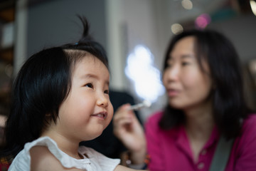 Fototapeta premium Selective focus of Asian adorable 2 years old toddler looks up smiling and enjoy having breakfast with the mother, while mom holding spoon prepare to feed her, concept mother and toddler.