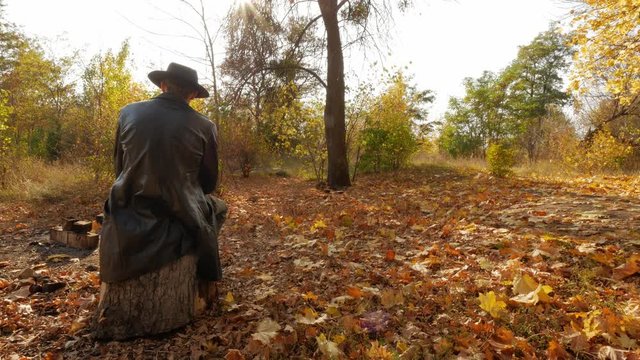 Mysterious man in a leather coat and hat sits in the autumn forest and waits for something.
