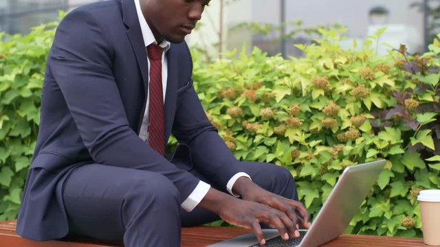 Young black male entrepreneur in formal clothes typing on laptop computer keyboard while working online on bench in park
